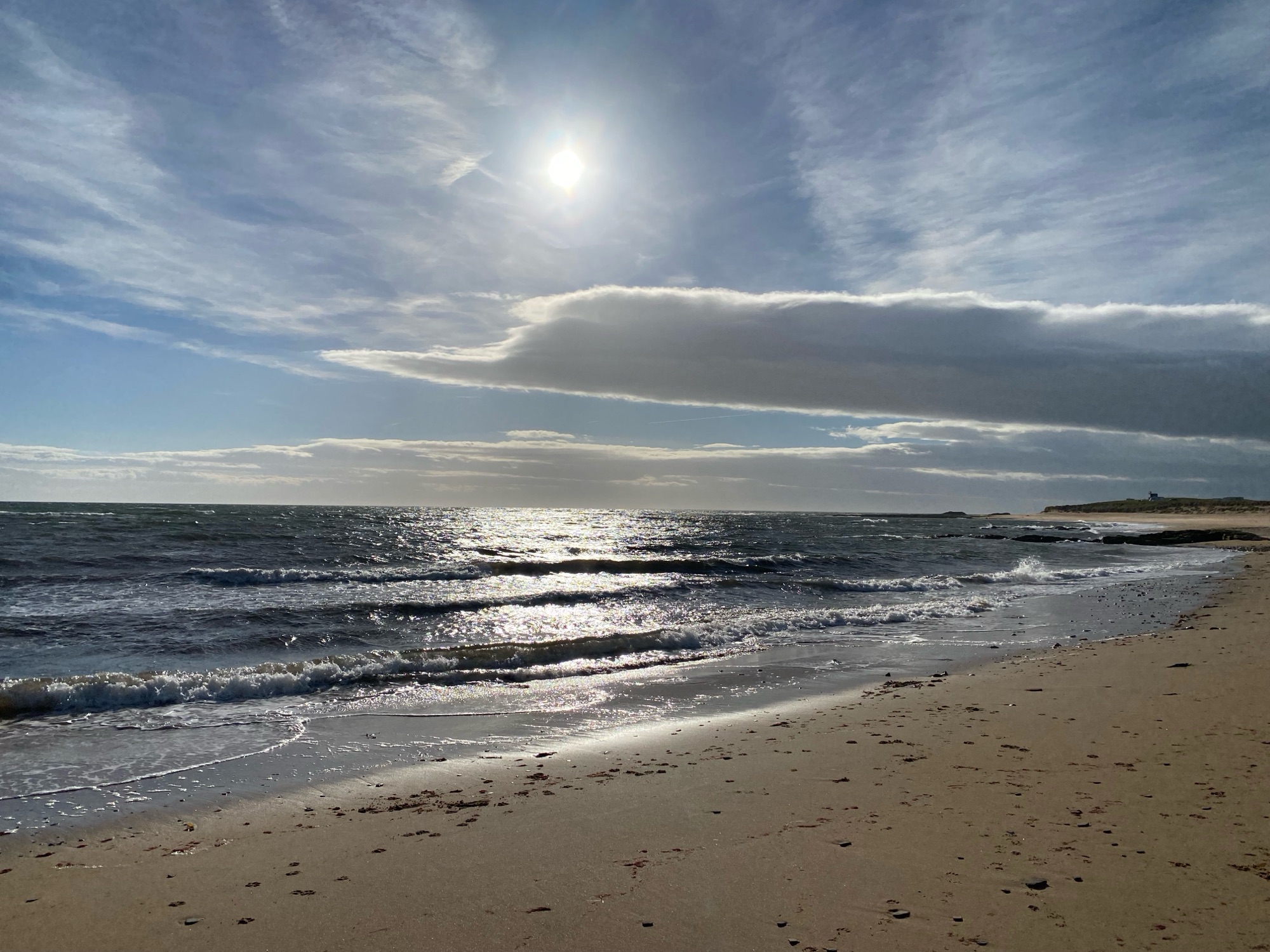 A weak sun in a bright cloudy blue sky over a beach with waves lapping the shore. The sun is silver on the water.
