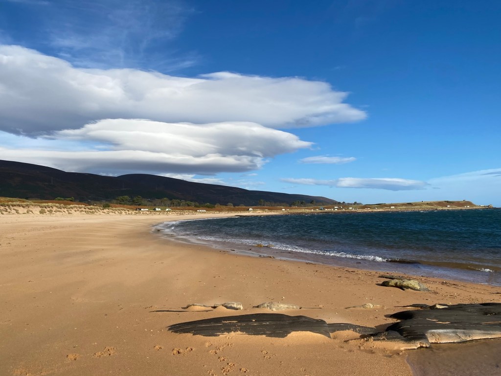 Large lenticular clouds hang in a very blue sky above a pinky sanded beach, where the waves are gently lapping the shore.